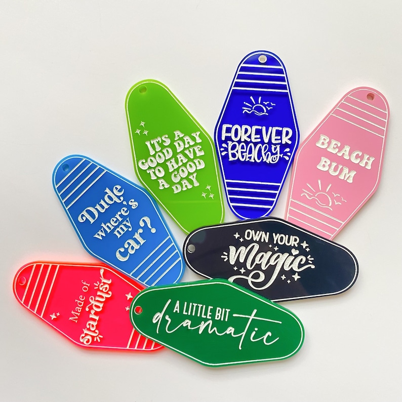Op de afbeelding: Assortiment van kleurrijke sleutelhangers in de vorm van motelsleutel met verschillende zinnen, waaronder "Dude where's my car?", "It's a good day to have a good day", "Forever Beachy", "Beach Bum", "Own Your Magic" en "A Little Bit Dramatic".