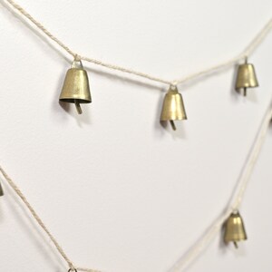 May include: A string of small gold bells hanging from a white twine. The bells are evenly spaced and hang in a diagonal line.