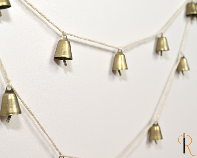 May include: A string of small gold bells hanging from a white twine. The bells are evenly spaced and hang in a diagonal line.