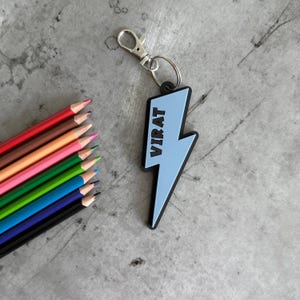 May include: A light blue lightning bolt-shaped keychain with the name "VIRAT" in white lettering. The keychain has a silver clasp and ring. A collection of colored pencils in various colors, including red, brown, pink, green, and blue, are arranged on the left.
