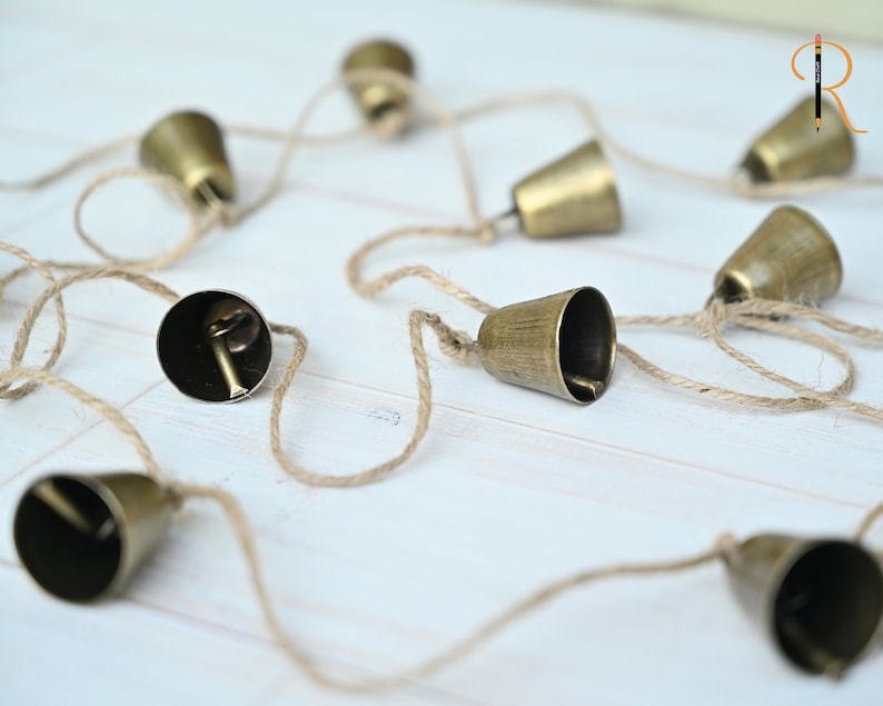 May include: A string of small, gold-colored bells hanging from a natural twine cord. The bells are arranged in a random pattern and are all facing different directions.