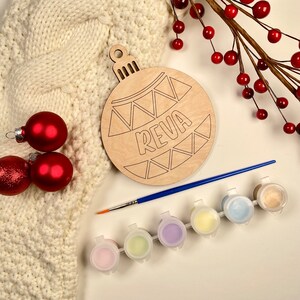 May include: A wooden Christmas ornament shaped like a bauble with the name "REVA" written on it. The ornament is surrounded by paint pots, a paintbrush, and red berries.