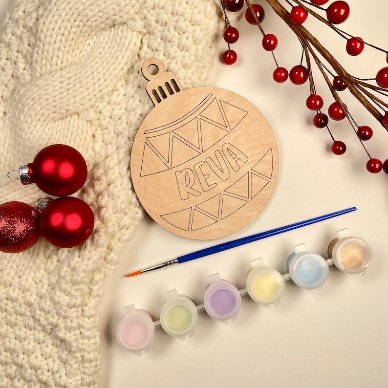 May include: A wooden Christmas ornament shaped like a bauble with the name "REVA" written on it. The ornament is surrounded by paint pots, a paintbrush, and red berries.