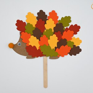 May include: A brown paper hedgehog with a googly eye and an orange pom-pom nose. The hedgehog is covered in colorful paper leaves in shades of red, orange, yellow, green, and brown. The hedgehog is attached to a wooden stick.
