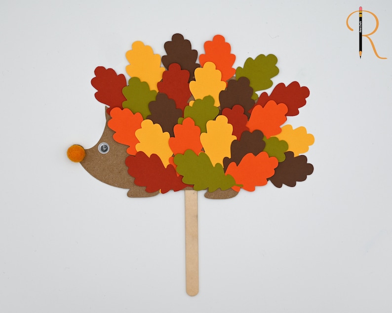 May include: A brown paper hedgehog with a googly eye and an orange pom-pom nose. The hedgehog is covered in colorful paper leaves in shades of red, orange, yellow, green, and brown. The hedgehog is attached to a wooden stick.