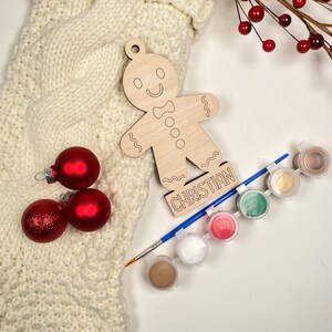 May include: Wooden gingerbread person ornament with the name Christian written below. The ornament is surrounded by red and white Christmas ornaments and paint pots with a paintbrush.