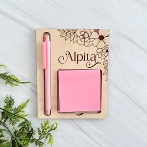May include: A wooden desk organiser with a pink pen and a pink sticky note pad. The organiser has the name "Alpita" engraved on it, along with a floral design. The pen is placed in a slot, and the notepad is in a recessed area.