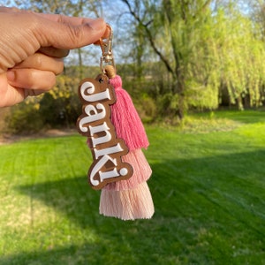 May include: A wooden keychain with the name "Janki" in white lettering. The keychain features a pink and light pink tassel.