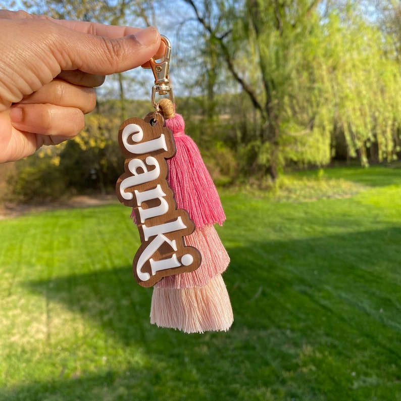 May include: A wooden keychain with the name "Janki" in white lettering. The keychain features a pink and light pink tassel.