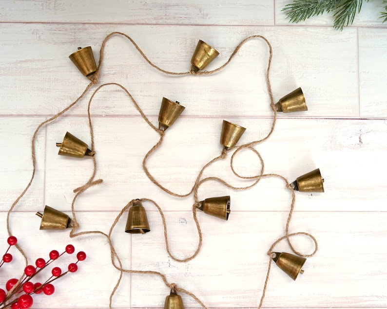 May include: A string of gold-colored bells hanging from a natural twine rope. The bells are arranged in a random pattern and are hanging on a white wooden background.