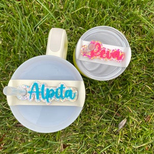 Può includere: Due bicchieri bianchi con coperchi. Un coperchio ha un'etichetta con il nome in glitter turchese che dice "Alpita" e l'altro coperchio ha un'etichetta con il nome in glitter rosa che dice "Leann".
