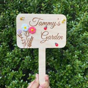 Pu&ograve; includere: Un cartello da giardino in legno con il testo "Tammy's Garden" in corsivo. Il cartello &egrave; decorato con piccoli fiori, coccinelle e api.