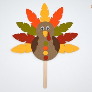 May include: A paper craft turkey with a brown body, orange, yellow, red, and green feathers, and googly eyes. The turkey is attached to a wooden stick.