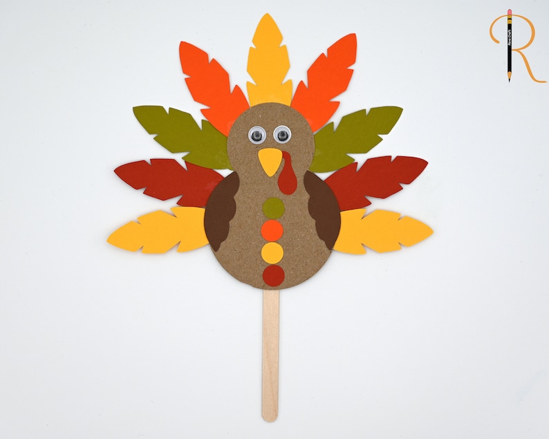 May include: A paper craft turkey with a brown body, orange, yellow, red, and green feathers, and googly eyes. The turkey is attached to a wooden stick.