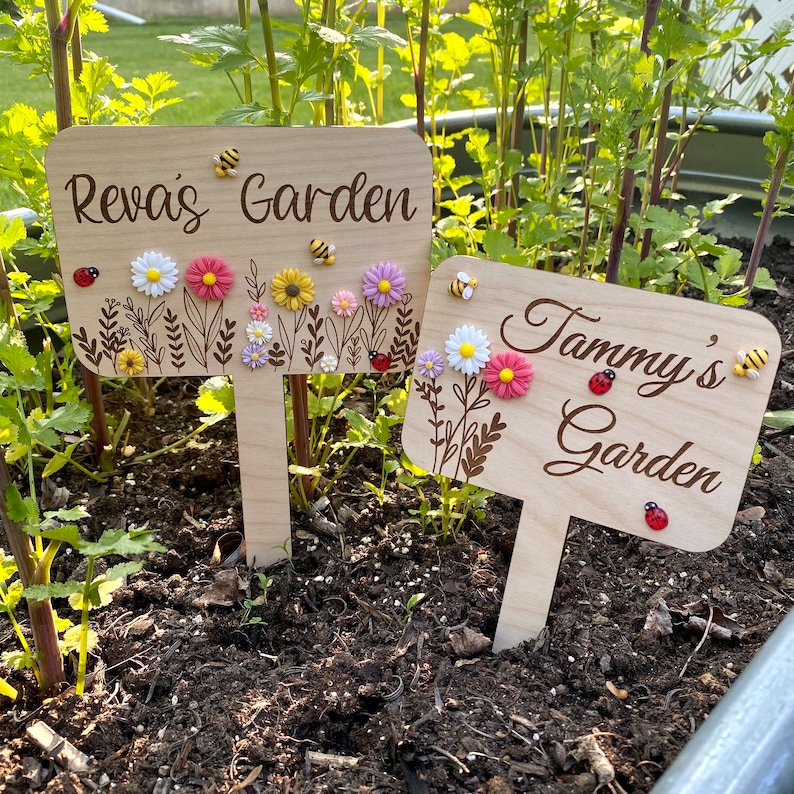 Pu&ograve; includere: Due cartelli da giardino in legno con i nomi "Reva's Garden" e "Tammy's Garden" incisi su di essi. I cartelli sono decorati con piccoli fiori e api.