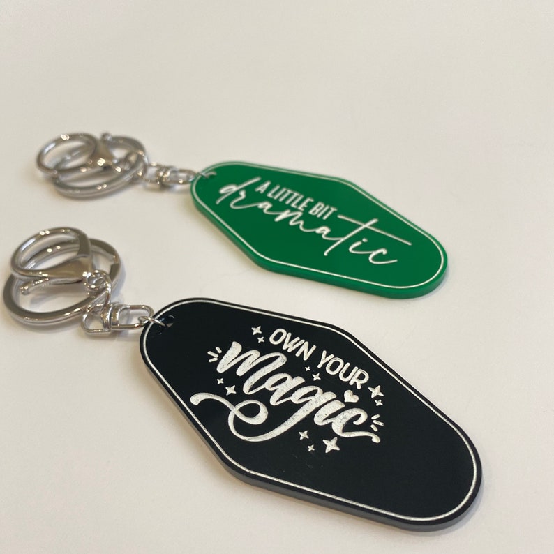 Op de afbeelding: Twee sleutelhangers met zwarte en groene motel-sleutel-vormige tags. De groene sleutelhanger heeft de tekst "A little bit dramatic" in witte schrijfletters. De zwarte sleutelhanger heeft de tekst "Own your magic" in witte schrijfletters met sterren en een hart.
