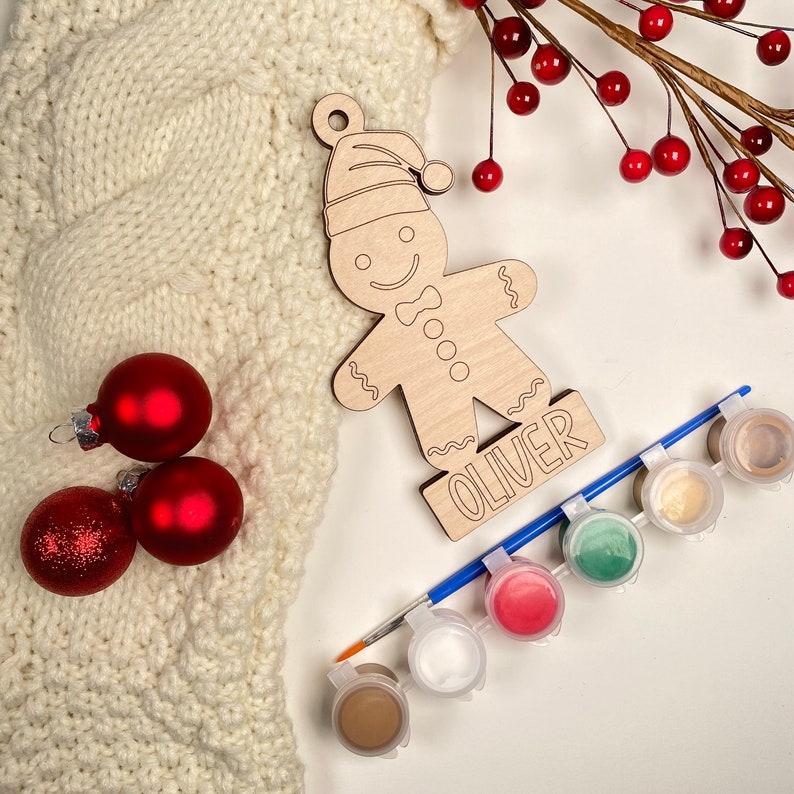 May include: Unpainted wooden gingerbread man ornament with the name "Oliver" written on the base. The ornament is surrounded by red and white Christmas ornaments, a paintbrush, and six small jars of paint in various colors.