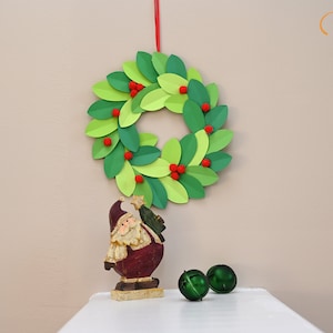 May include: A green paper wreath with red pom-poms hangs on a wall. A small Santa figurine and two green jingle bells are on a white surface below the wreath.