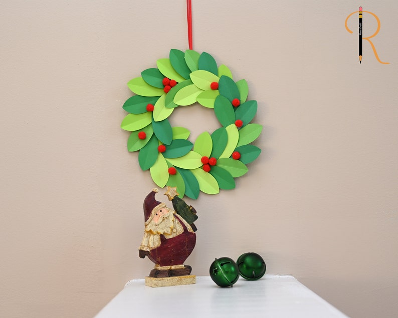 May include: A green paper wreath with red pom-poms hangs on a wall. A small Santa figurine and two green jingle bells are on a white surface below the wreath.