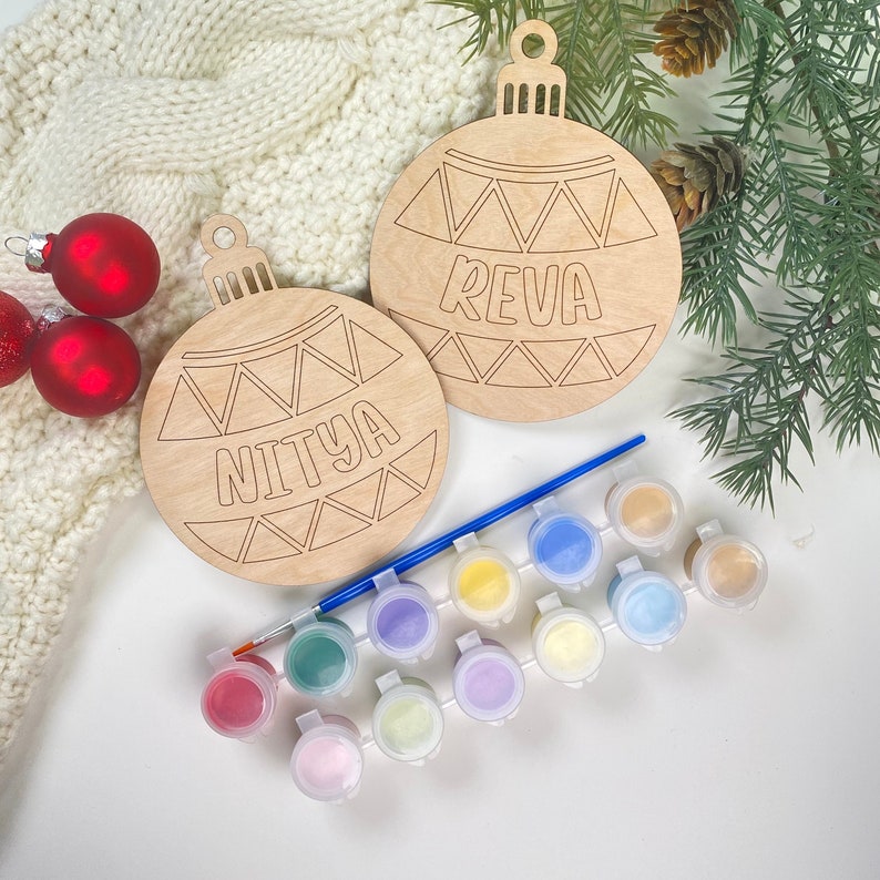 May include: Two unfinished wooden Christmas ornament shapes with the names "Reva" and "Nitya" written on them. The ornaments are surrounded by paint pots and a paintbrush.