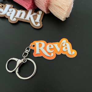 May include: Orange and white acrylic keyring with the name "Reva" in a retro font. The keyring has a silver metal ring.