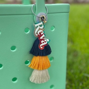 May include: A green tote bag with a personalised acrylic keychain featuring the name "Reva" in white letters on a colourful background. The keychain has a multi-coloured tassel.