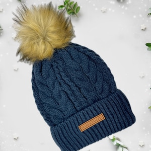 May include: A navy blue cable knit beanie with a faux fur pom pom and a brown leather patch that says "Peaks & Valleys".
