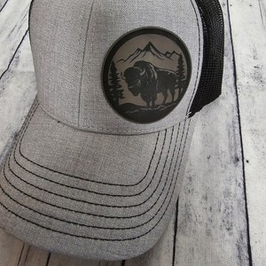 May include: A gray and black trucker hat with a black leather patch featuring a silhouette of a buffalo in front of a mountain range.