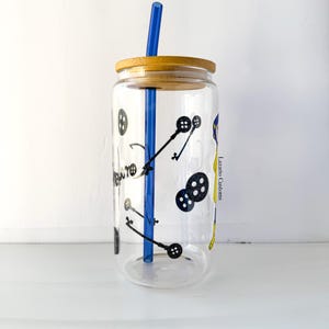 May include: A clear glass tumbler with a bamboo lid and a blue straw. The tumbler is decorated with black illustrations of keys and buttons. The text "Lazaro Customs" is visible on the side of the glass.