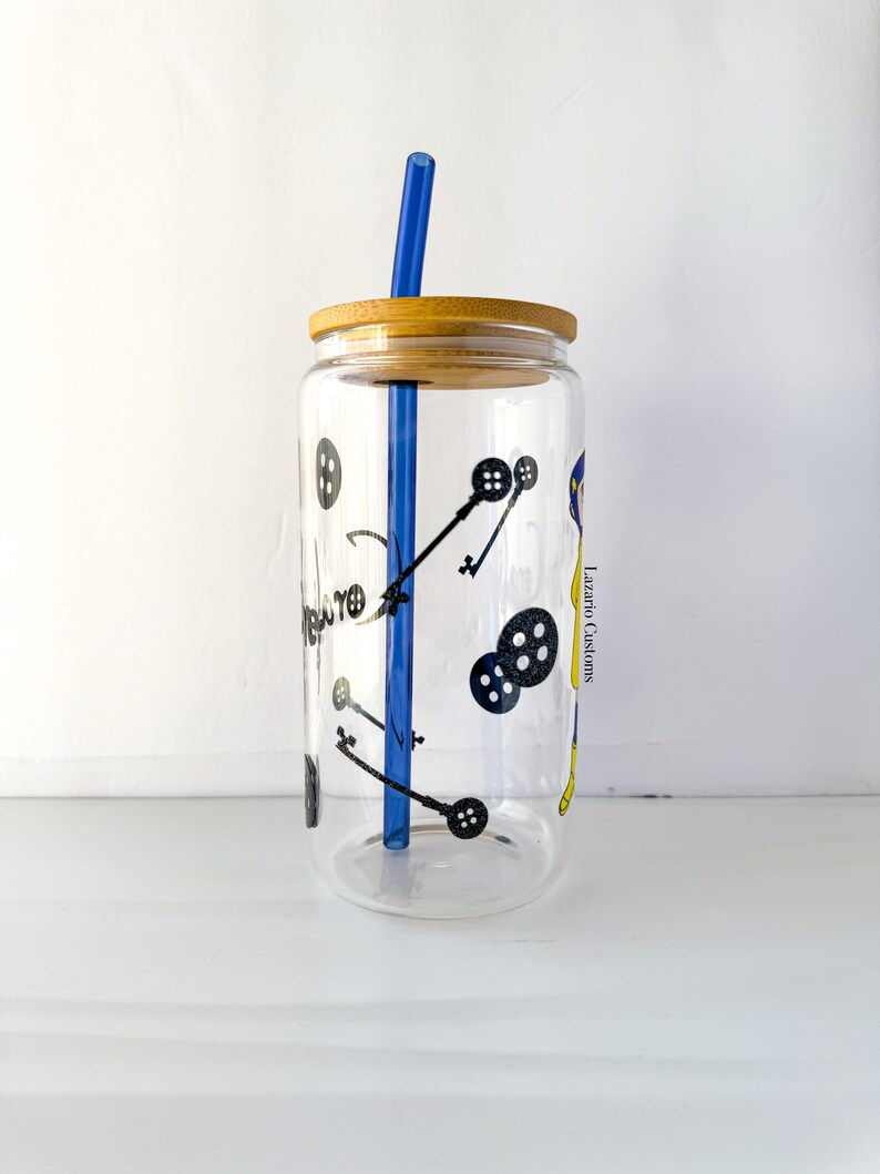 May include: A clear glass tumbler with a bamboo lid and a blue straw. The tumbler is decorated with black illustrations of keys and buttons. The text "Lazaro Customs" is visible on the side of the glass.