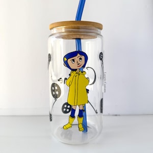 May include: A clear glass tumbler with a bamboo lid and blue straw. The tumbler features a cartoon illustration of a character in a yellow raincoat and boots. Black button graphics are scattered around the glass.
