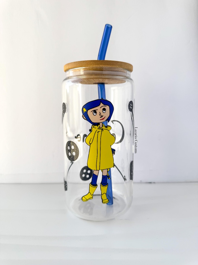 May include: A clear glass tumbler with a bamboo lid and blue straw. The tumbler features a cartoon illustration of a character in a yellow raincoat and boots. Black button graphics are scattered around the glass.