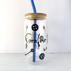 May include: A clear glass tumbler with a bamboo lid and a blue straw. The tumbler is decorated with black button and key graphics, and the name "Coraline" in a stylized font. The tumbler is designed for cold beverages.