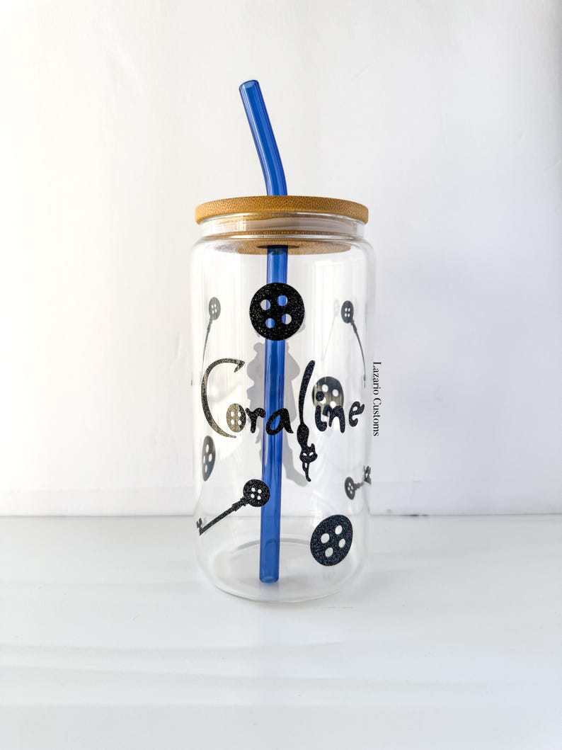 May include: A clear glass tumbler with a bamboo lid and a blue straw. The tumbler is decorated with black button and key graphics, and the name "Coraline" in a stylized font. The tumbler is designed for cold beverages.