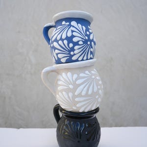 May include: Three stacked ceramic mugs. The top mug is blue with white floral designs, the middle mug is beige with white floral designs, and the bottom mug is black with carved floral designs. Each mug has a handle.