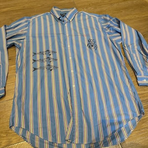 May include: Blue striped button-down shirt with vertical yellow and red stripes. The shirt has embroidered fish and a crest. Long-sleeved with a button-down collar.