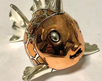 May include: A small silver and copper fish figurine with a textured body and large eyes. The fish has a fin on its back and a tail fin.