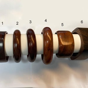 Könnte beinhalten: Eine Sammlung von sechs Holzarmreifen in verschiedenen Designs und Brauntönen. Die Armreifen werden auf einem weißen zylindrischen Ständer präsentiert. Die Holzmaserung ist auf jedem Stück sichtbar und zeigt die natürliche Schönheit des Materials.