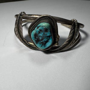 May include: A silver cuff bracelet with a large, oval turquoise stone at the center. The stone is a vibrant blue-green with dark matrix patterns. The bracelet features leaf-shaped accents on either side of the stone.