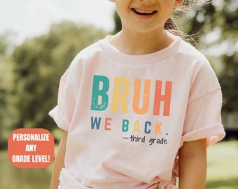 Op de afbeelding: Een roze T-shirt met de tekst "BRUH WE BACK. -third grade" in kleurrijke letters. Het shirt wordt gedragen door een kind.
