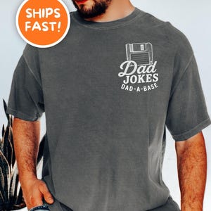 May include: A charcoal gray short-sleeve t-shirt featuring a white graphic on the left chest. The graphic depicts a floppy disk icon above the text "Dad JOKES" and "DAD-A-BASE" in a clean, sans-serif font. The shirt has a crew neckline.