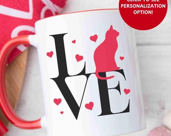 May include: White ceramic mug with a red rim and handle. The mug features a black graphic of the word "LOVE" with a pink cat silhouette inside the letter "E". There are small pink hearts scattered around the graphic.