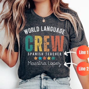 May include: Dark heather grey t-shirt with the text "WORLD LANGUAGE CREW" in white and orange, and "SPANISH TEACHER" in white. Below is "Maestra Lopez" in white. Colorful stars are below the text.