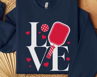 May include: A navy blue sweatshirt with a white graphic of the word "LOVE" with a red and white pickleball paddle and ball in the middle of the "O".