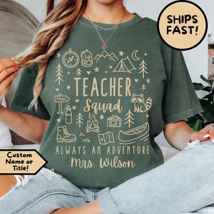 May include: A moss green short-sleeve t-shirt featuring a cream-colored graphic print with the text "TEACHER SQUAD", "ALWAYS AN ADVENTURE", and "Mrs. Wilson". The design includes icons of a compass, tent, trees, raccoon, camera, canoe, and hiking boots.
