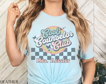 May include: A light blue t-shirt with a retro-style graphic that says "Cool Counselor Club" with a rainbow and butterflies. The text "Mrs. Ressler" is below the graphic.