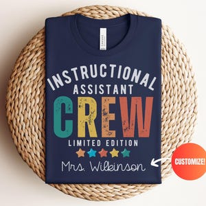 May include: Navy blue t-shirt featuring the text "INSTRUCTIONAL ASSISTANT CREW" in white, teal, yellow, and orange. The shirt also includes the text "LIMITED EDITION" and "Mrs. Wilkinson" with colorful stars. The t-shirt is folded on a woven mat.