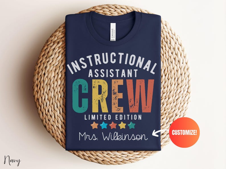 May include: Navy blue t-shirt featuring the text "INSTRUCTIONAL ASSISTANT CREW" in white, teal, yellow, and orange. The shirt also includes the text "LIMITED EDITION" and "Mrs. Wilkinson" with colorful stars. The t-shirt is folded on a woven mat.