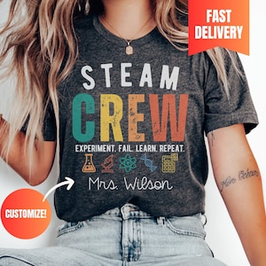 May include: A dark heather grey t-shirt featuring the white text STEAM and the multi-colored distressed text CREW. Below is the text EXPERIMENT. FAIL. LEARN. REPEAT. with icons of a flask, microscope, atom, DNA strand, and calculator, plus Mrs. Wilson.