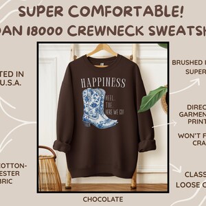 May include: A chocolate-coloured Gildan 18000 crewneck sweatshirt featuring a blue and white floral cowboy boot design. The word "HAPPINESS" is above the boot graphic, with "HEEL TOE HERE WE GO!" below. Made from a 50/50 cotton-polyester blend.